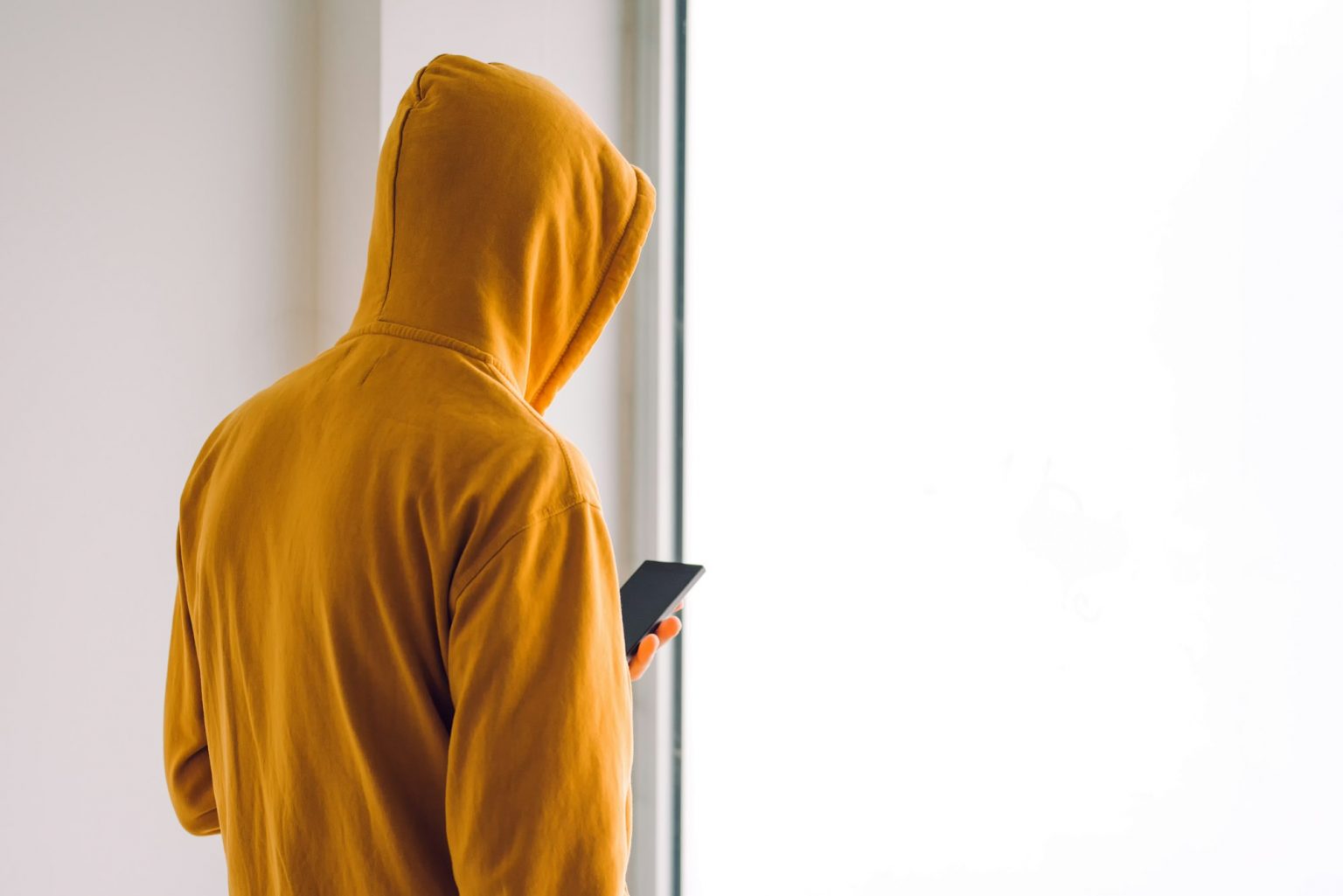 Les arnaques amoureuses sur Internet : décryptage et solutions Man with hoodie and mobile phone standing by the window